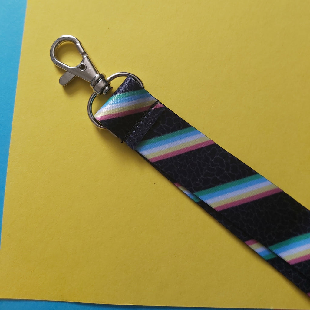 A close-up of a lanyard with a silver lobster clasp, featuring a black textured background and diagonal rainbow-colored stripes in pink, blue, green, and yellow.