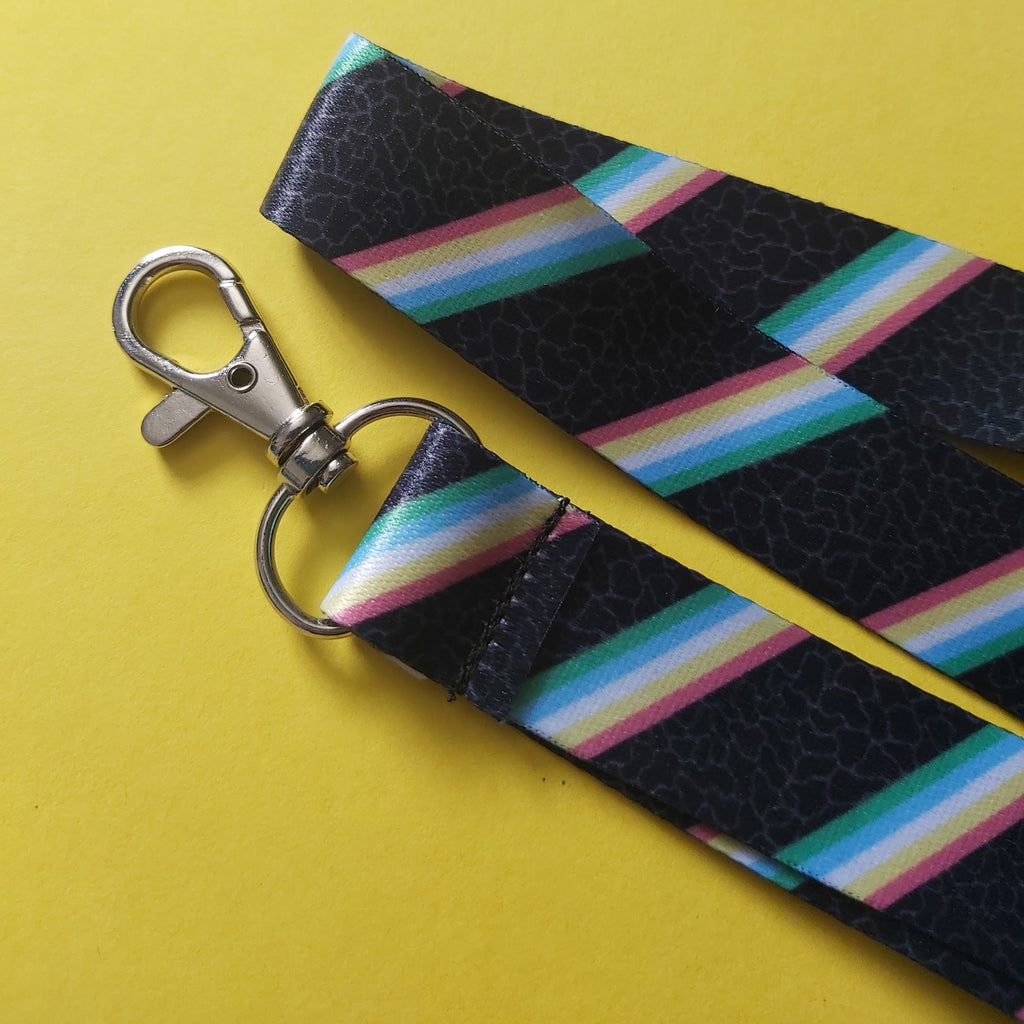 A close-up of a fabric lanyard strap with a silver metal swivel clasp, featuring a black textured background with diagonal rainbow stripes, set against a solid yellow background.