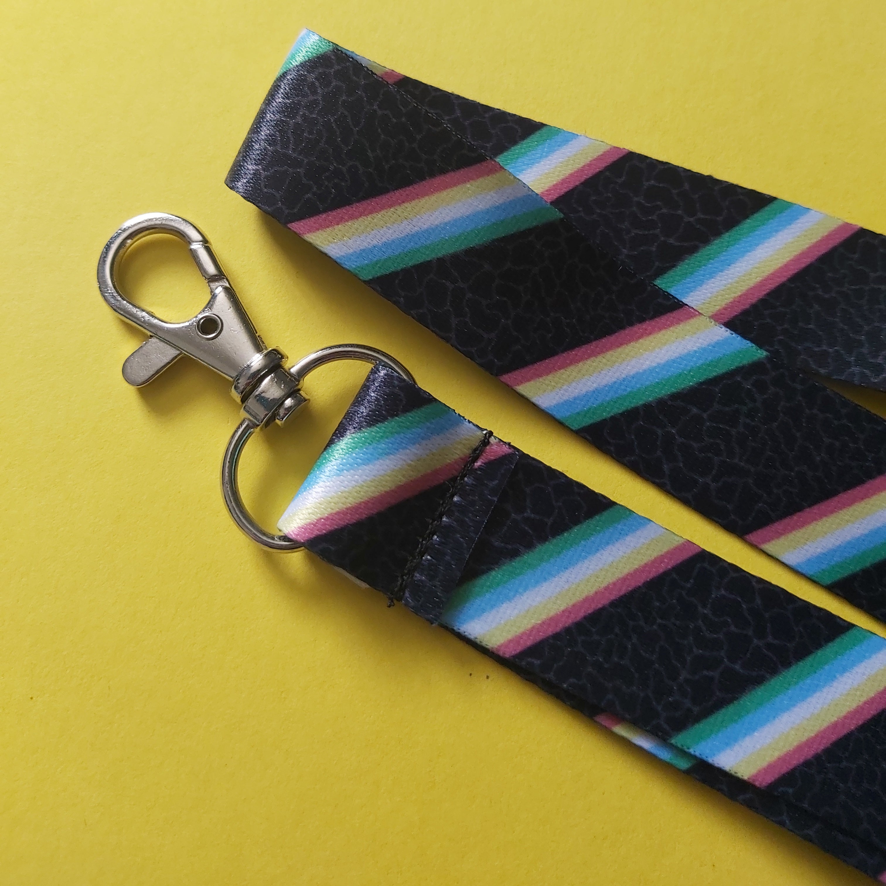 A close-up of a fabric lanyard strap with a silver metal swivel clasp, featuring a black textured background with diagonal rainbow stripes, set against a solid yellow background.