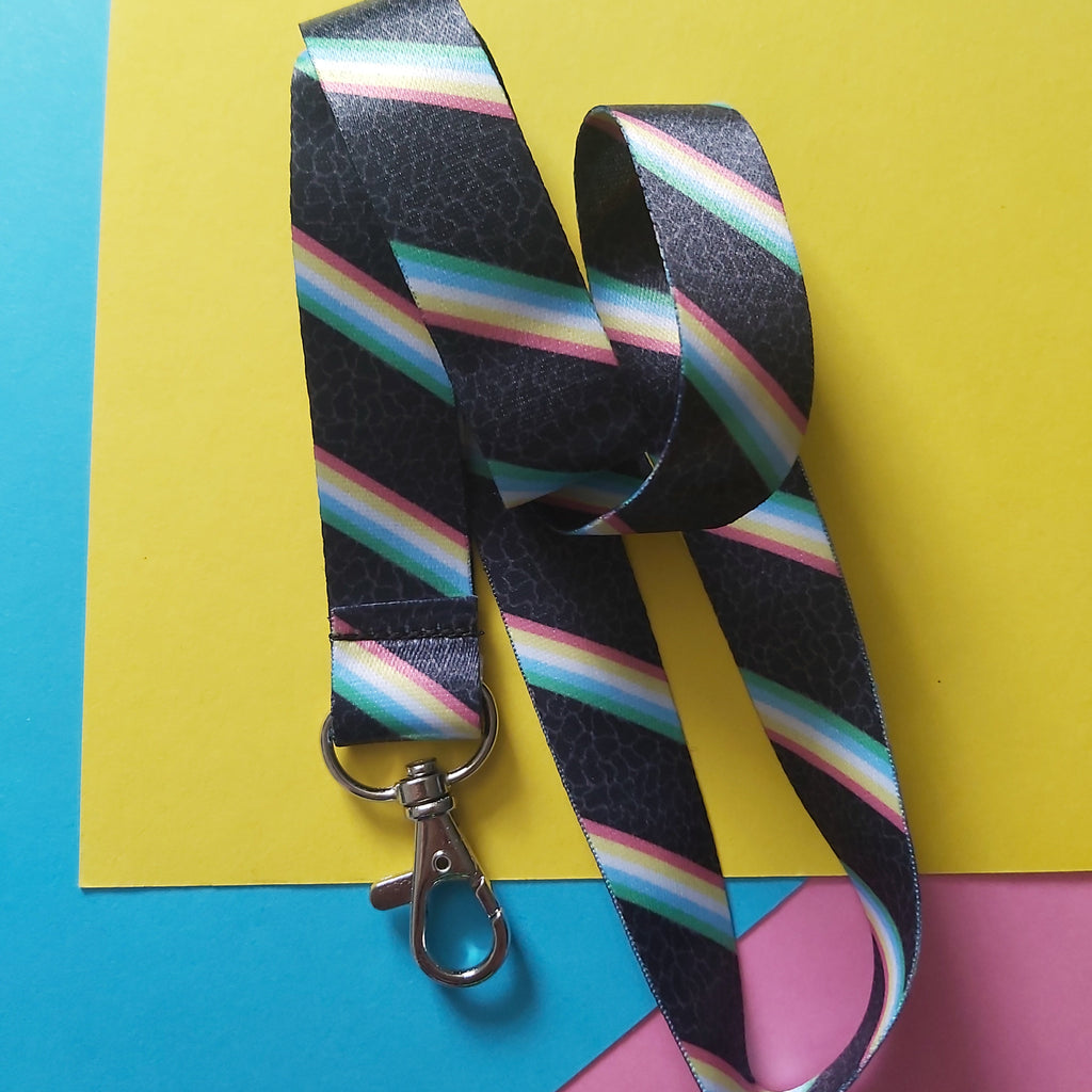 A black lanyard strap with a repeating diagonal rainbow stripe pattern and a silver metal lobster clasp, displayed on a colourful background of yellow, blue, and pink paper.