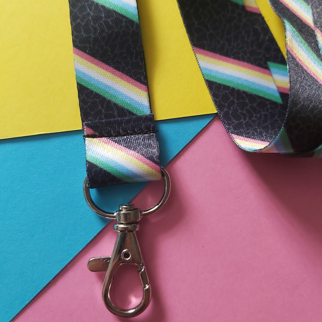 A close-up of a black lanyard with diagonal rainbow stripes and a silver metal lobster clasp, laid on a colorful background of yellow, blue, and pink paper.