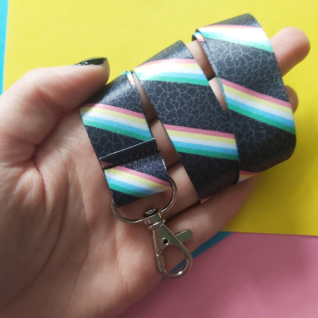 A hand holding a black lanyard strap with a colourful rainbow stripe pattern and a silver metal lobster clasp, set against a bright yellow and pink background.
