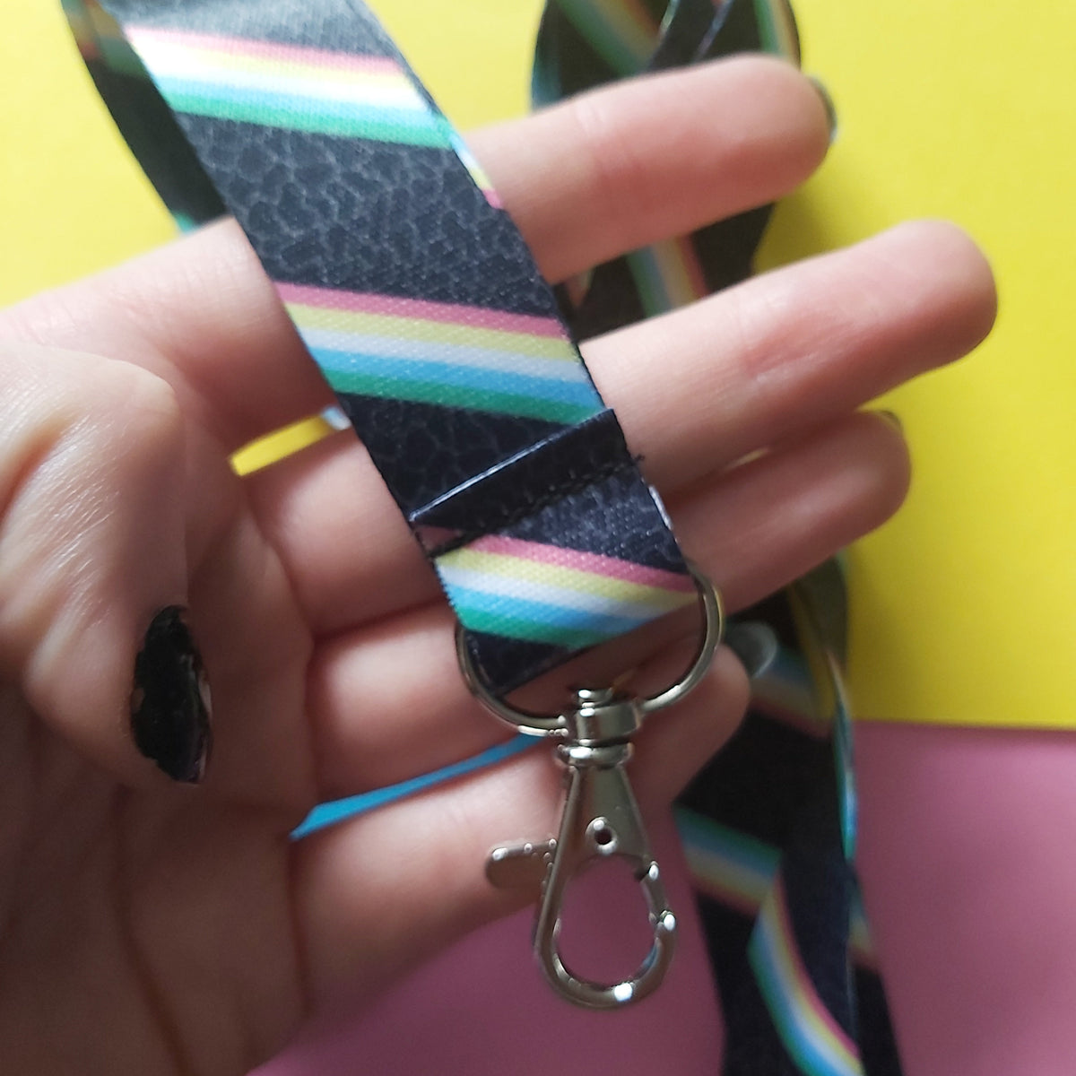 A close-up of a hand holding a lanyard strap with a colourful rainbow stripe pattern and a silver metal lobster clasp, set against a split yellow and pink background.