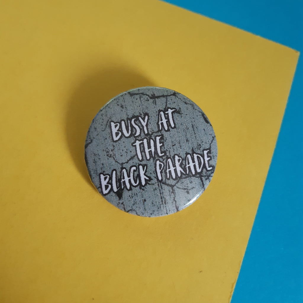 A round pin badge with a distressed grey background and white text reading 'BUSY AT THE BLACK PARADE', placed on a yellow sheet of paper over a blue background.