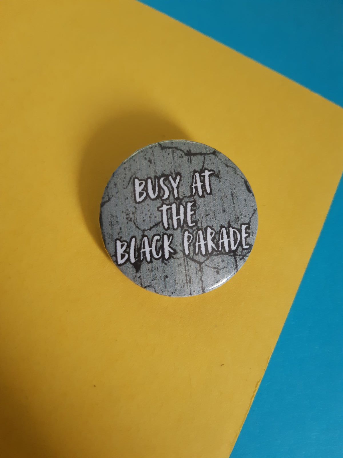 A round pin badge with a distressed grey and black textured background and white text reading 'BUSY AT THE BLACK PARADE', placed on a yellow paper background with a blue corner visible.