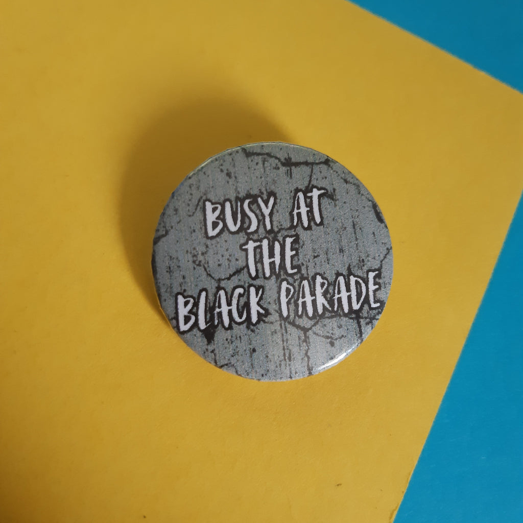 A round pin badge with a distressed gray and black textured background and white text reading 'BUSY AT THE BLACK PARADE', placed on a yellow paper background with a blue corner visible.