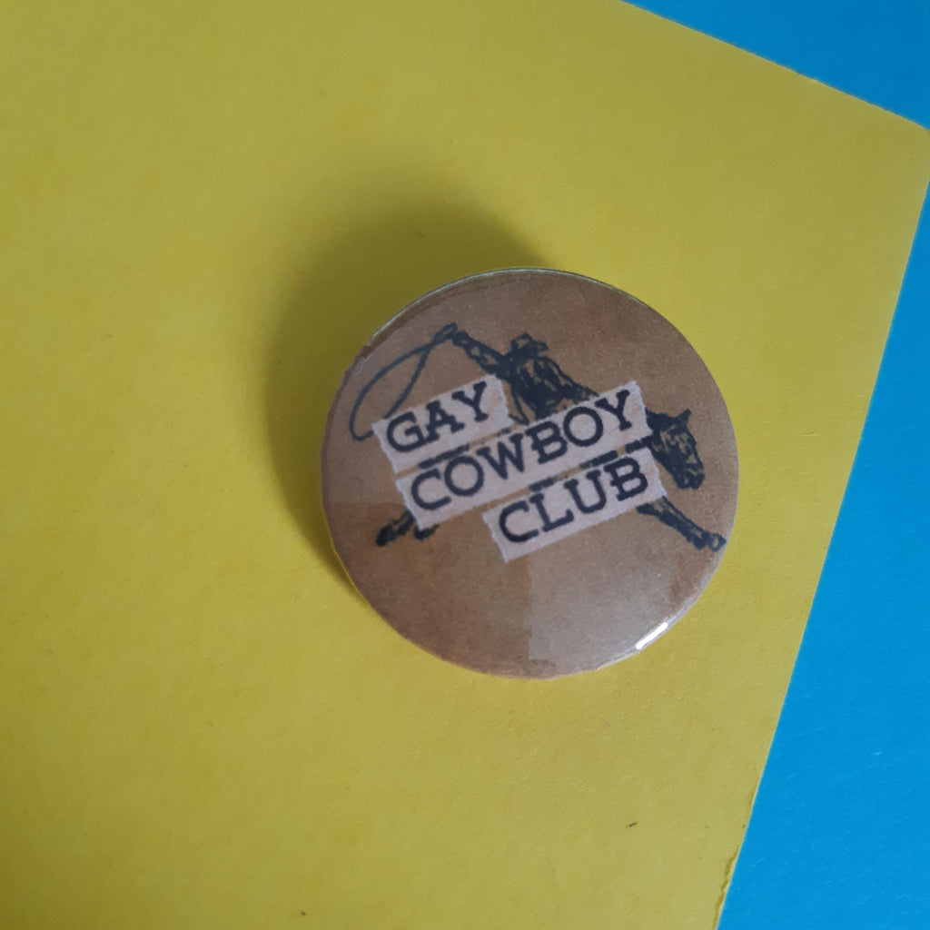 A round pinback button badge with a brown background and black western-themed graphic, featuring the text 'GAY COWBOY CLUB' in white lettering, placed on a yellow surface with a blue corner visible.