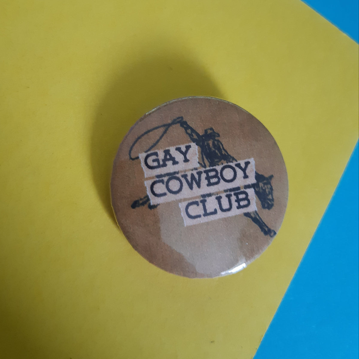 A round button badge with the text 'GAY COWBOY CLUB' and a cowboy illustration, placed on a yellow surface with a blue corner visible in the background.