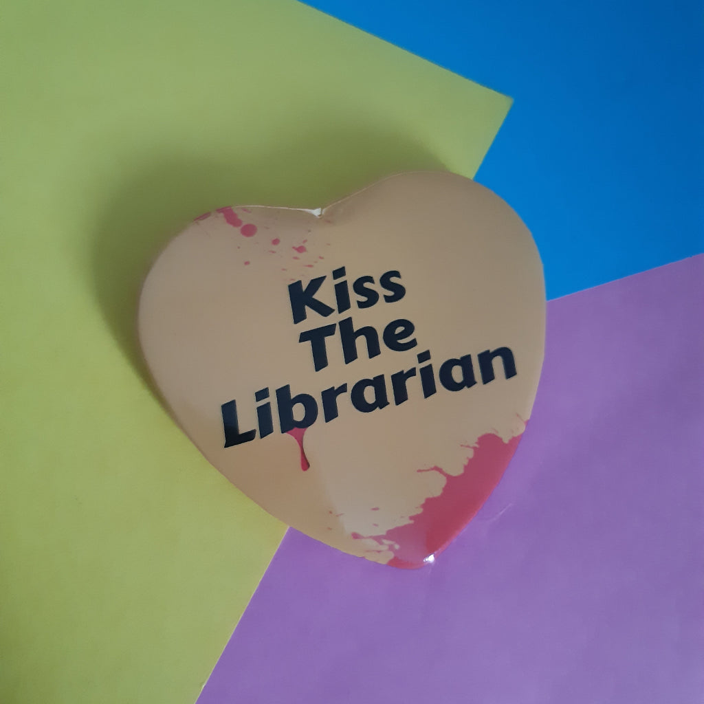 A yellow heart-shaped pinback button with the text 'Kiss The Librarian' in black, featuring red blood-splatter details, displayed on a background of green, blue, and purple paper.