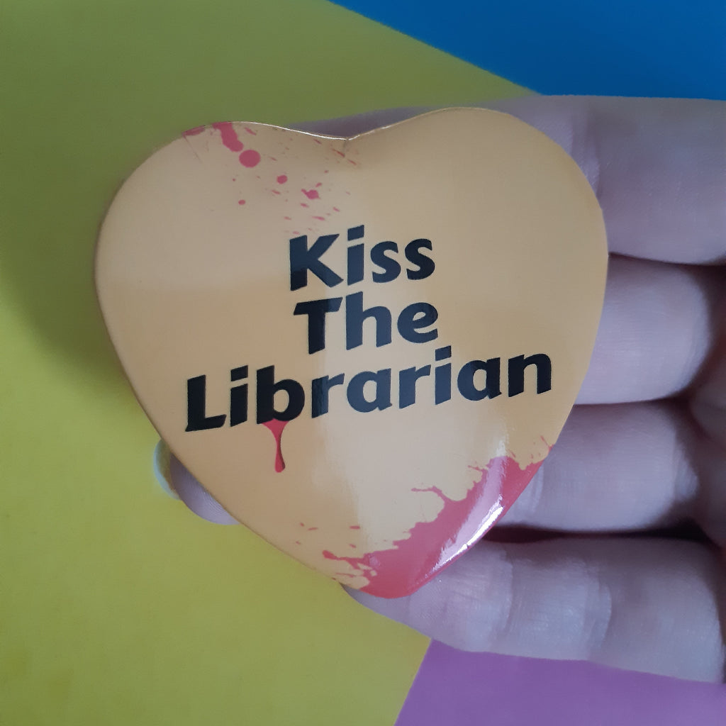 A hand holding a heart-shaped pinback button badge with the text 'Kiss The Librarian' in black, featuring red blood splatter details on a peach-colored background, set against a colourful background of yellow, blue, and pink paper.