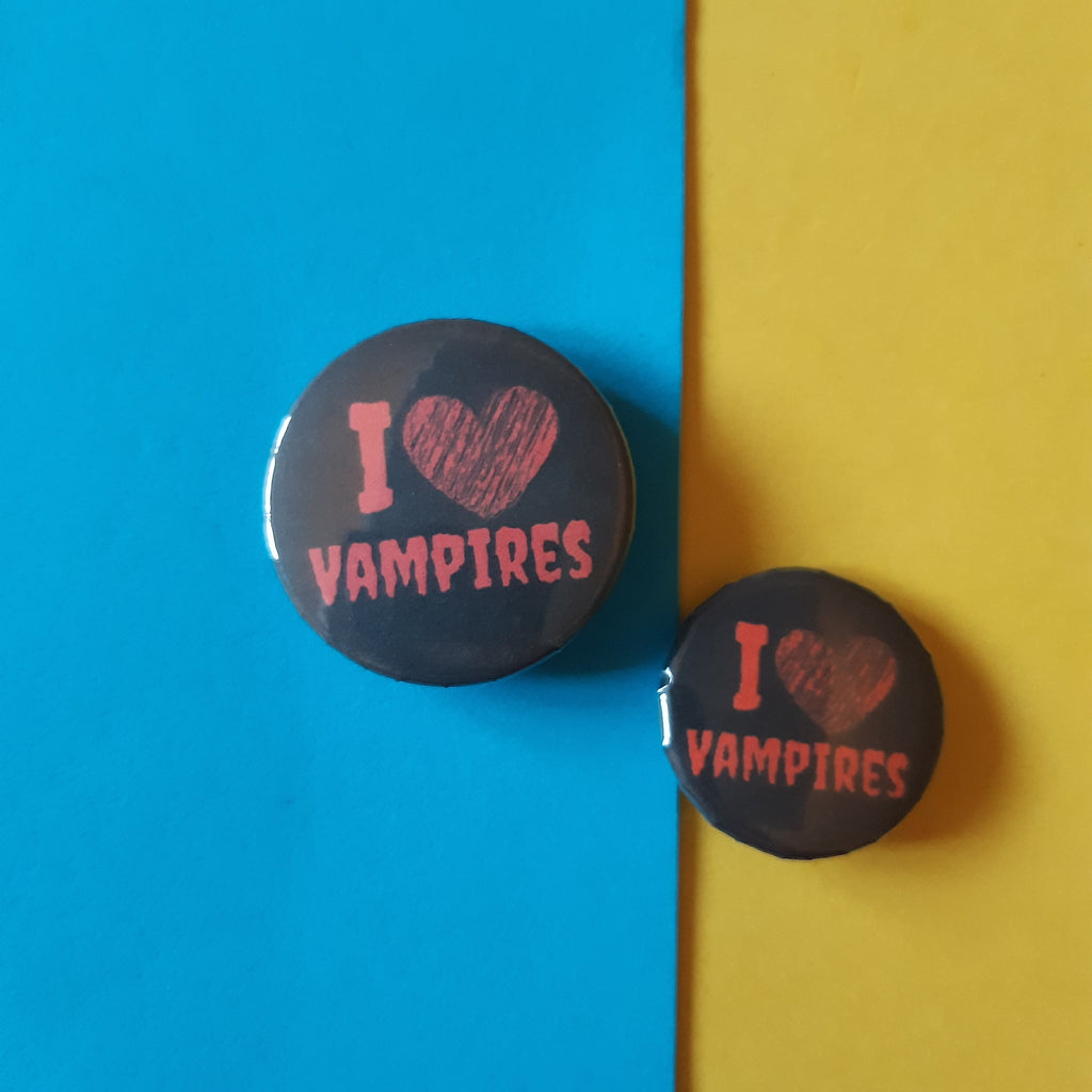 Two round black pin-back buttons with pink text reading 'I [heart] VAMPIRES' are displayed on a split blue and yellow background.