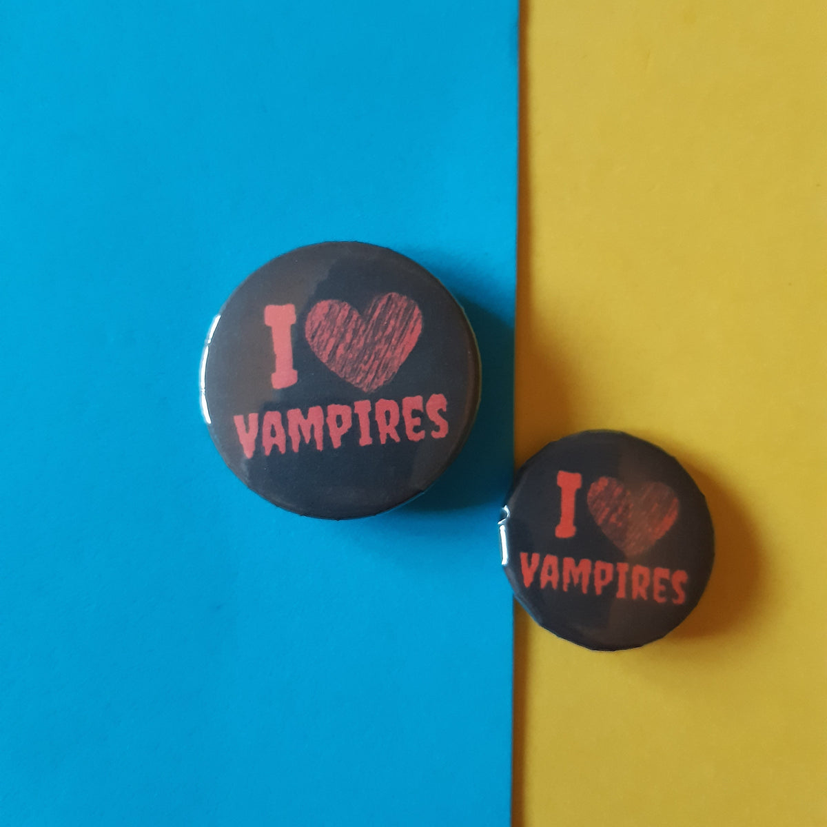 Two round black pin-back buttons with pink text reading 'I [heart] VAMPIRES' are displayed on a split blue and yellow background.