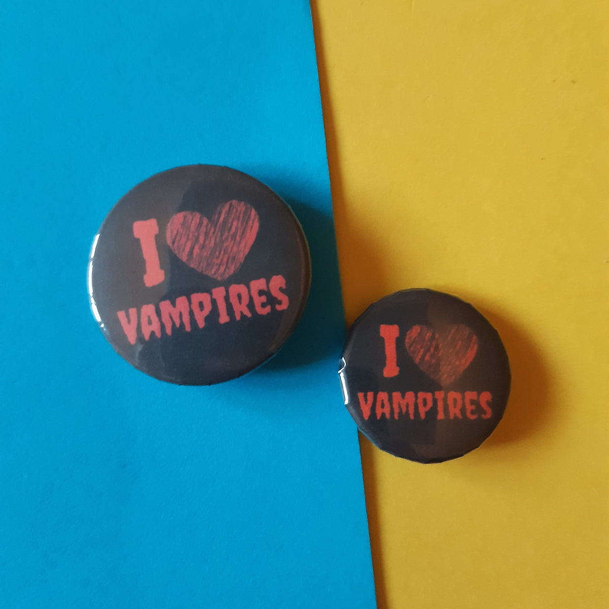Two round pin-back buttons with the text 'I [heart] VAMPIRES' in red on a dark background, displayed on a split blue and yellow paper backdrop.