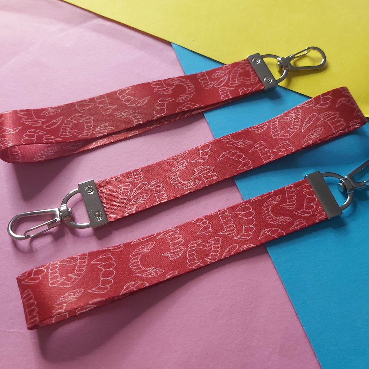 Three red fabric lanyards with a white patterned fang design and silver metal swivel clips, laid out diagonally on a colourful background of pink, blue, and yellow paper.