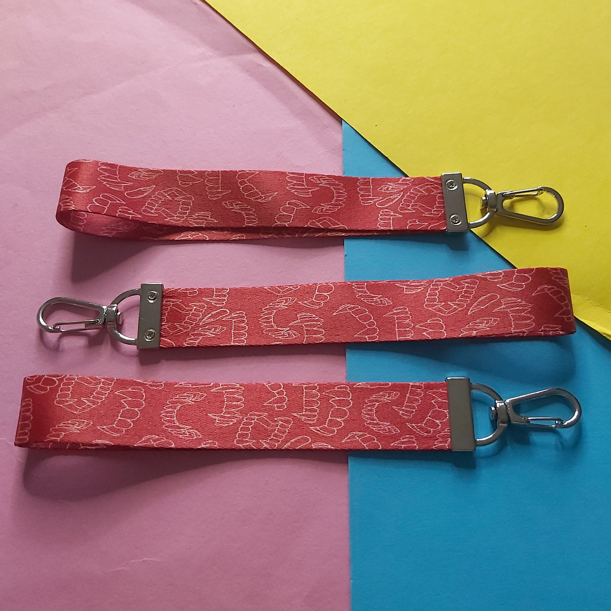 Three red fabric wristlets with a white patterned design and silver metal swivel clasp hardware, laid out on a background of pink, blue, and yellow paper.