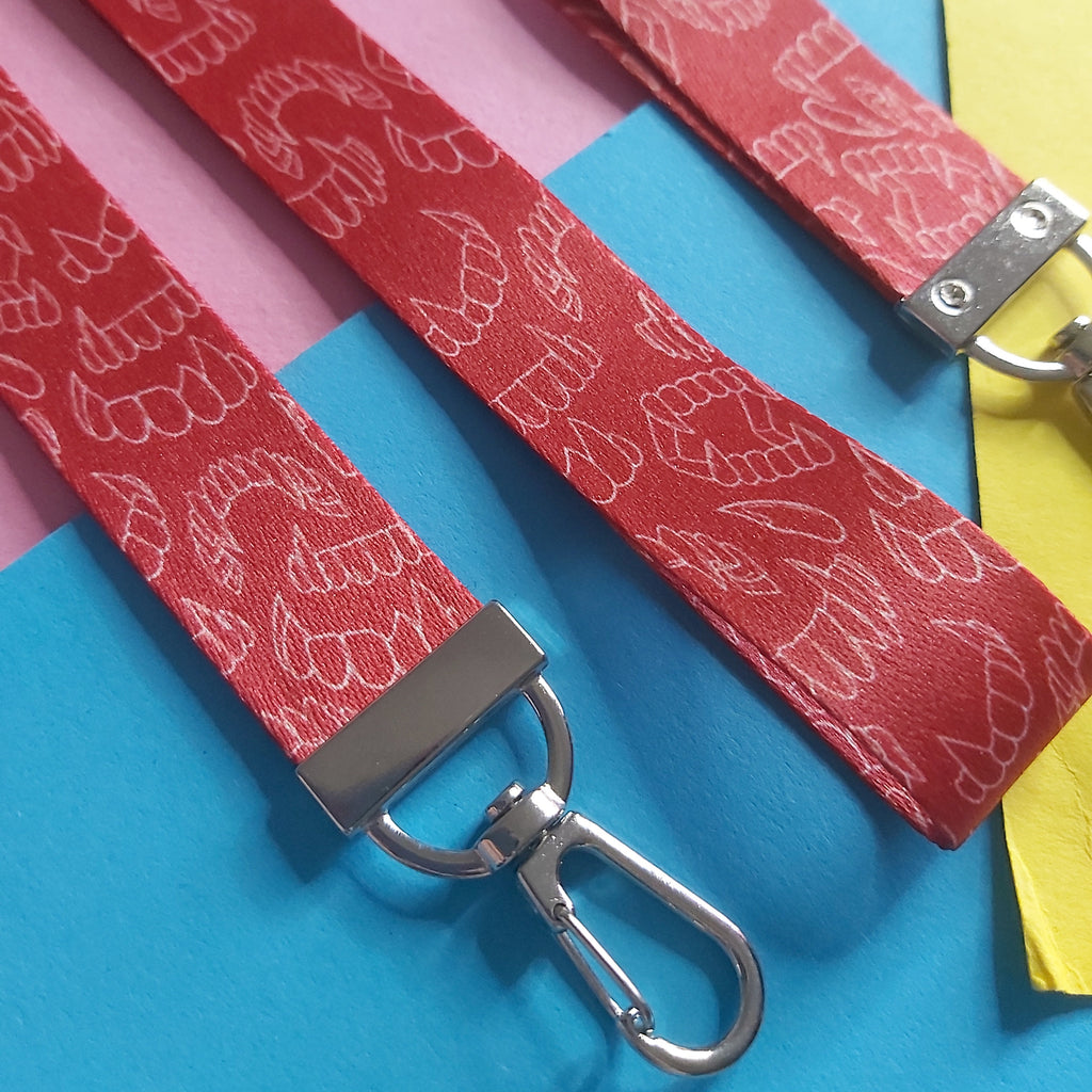 Red wristlet straps with a white line-art pattern of vampire fangs, featuring silver metal swivel clips, laid out on a colourful background of pink, blue, and yellow paper.