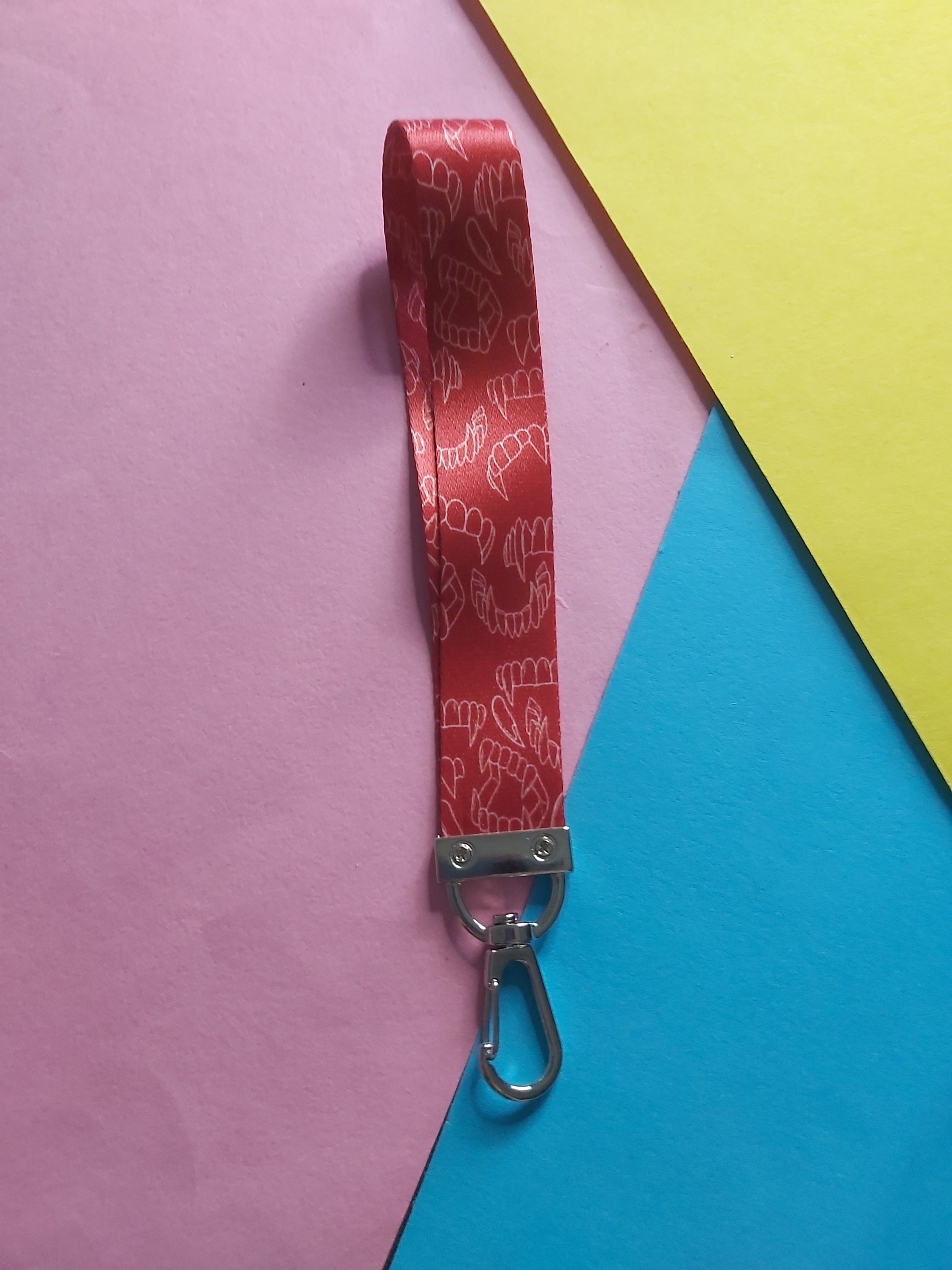 A red fabric wristlet strap with a repeating white fang pattern and a silver metal lobster clasp, laid diagonally on a colourful background of pink, yellow, and blue paper.