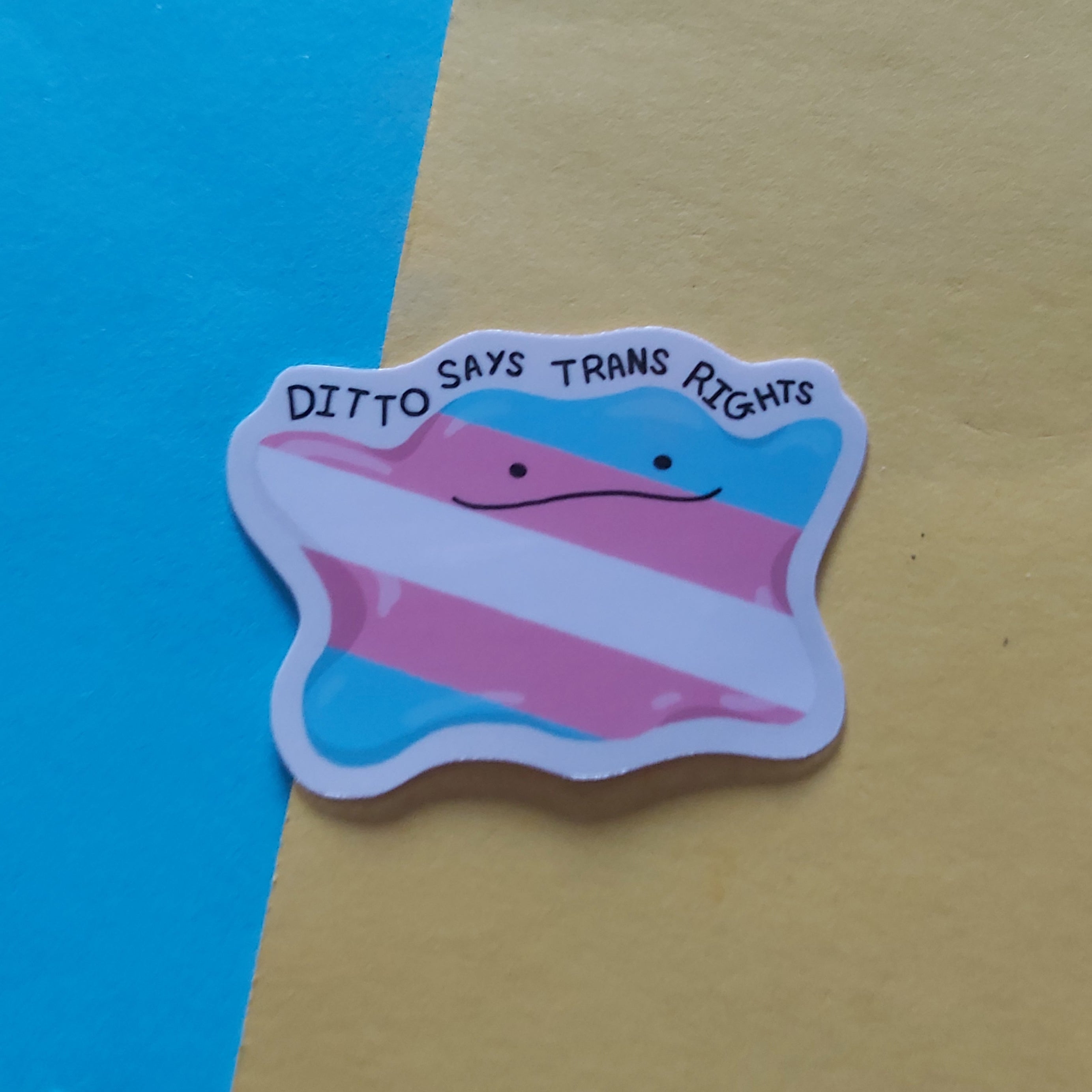 A die-cut sticker featuring a cartoon face with the text 'DITTO SAYS TRANS RIGHTS' over horizontal pink, white, and blue stripes, placed on a yellow sheet over a bright blue background.