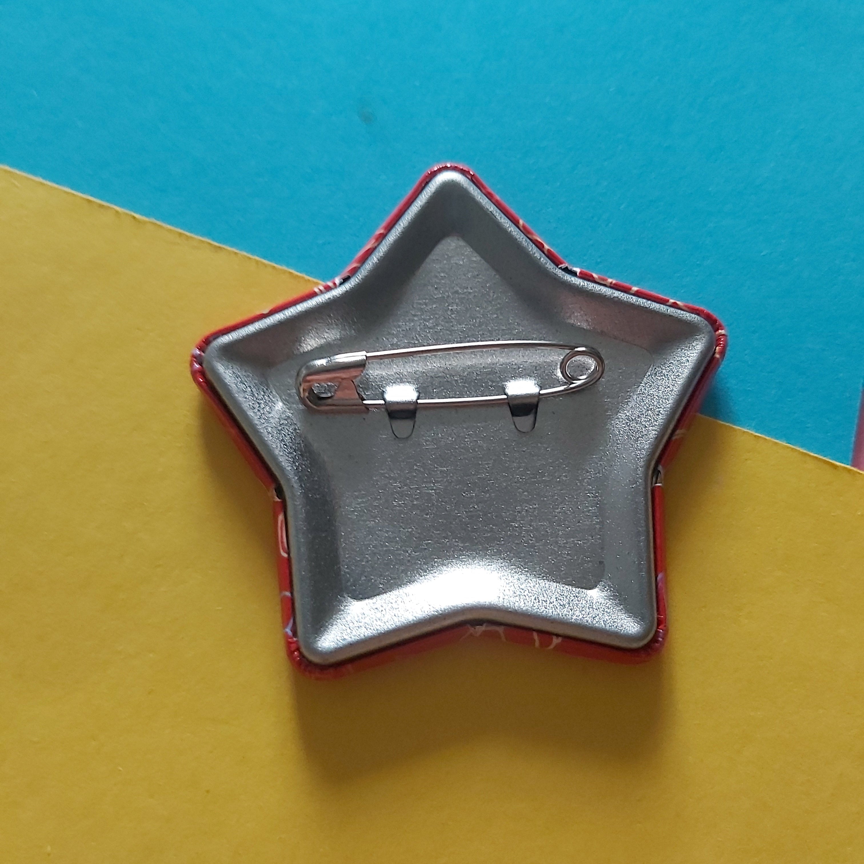 The back of a star-shaped pin badge with a silver metal safety pin clasp and a red border, shown on a split yellow and blue background.