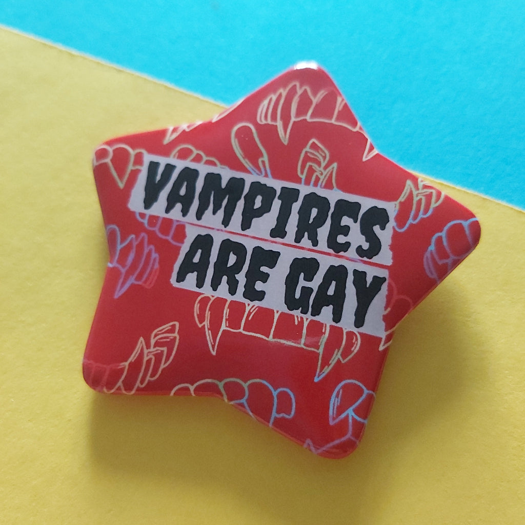 A red star-shaped pin with the text 'VAMPIRES ARE GAY' in black letters on a white banner, decorated with white and pink vampire illustrations, set against a split yellow and blue background.