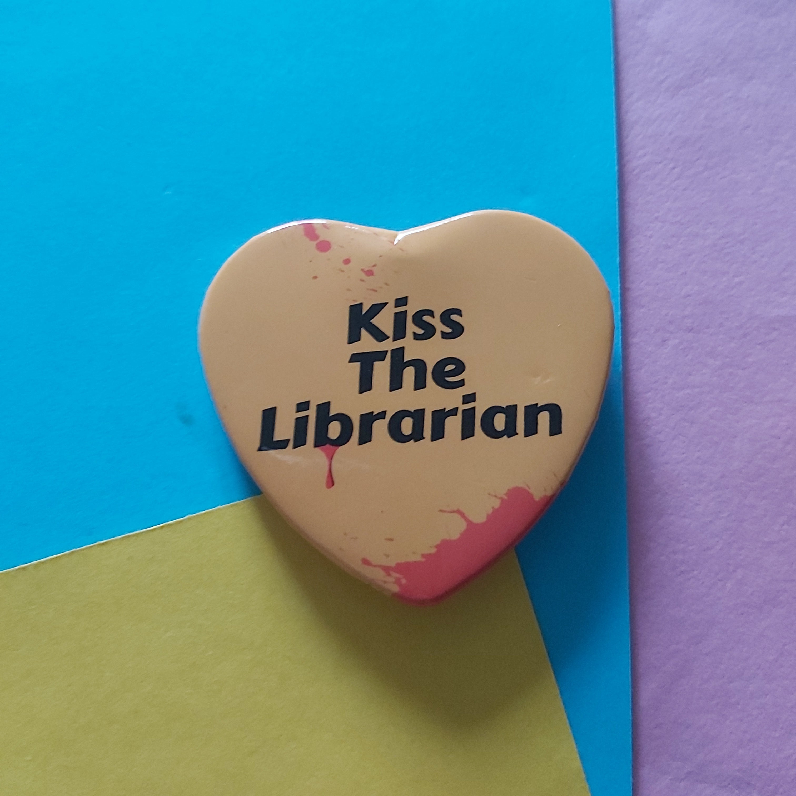 A heart-shaped pin badge with the text 'Kiss The Librarian' in black, decorated with red blood splatters, resting on a colourful background of blue, yellow, and purple paper.