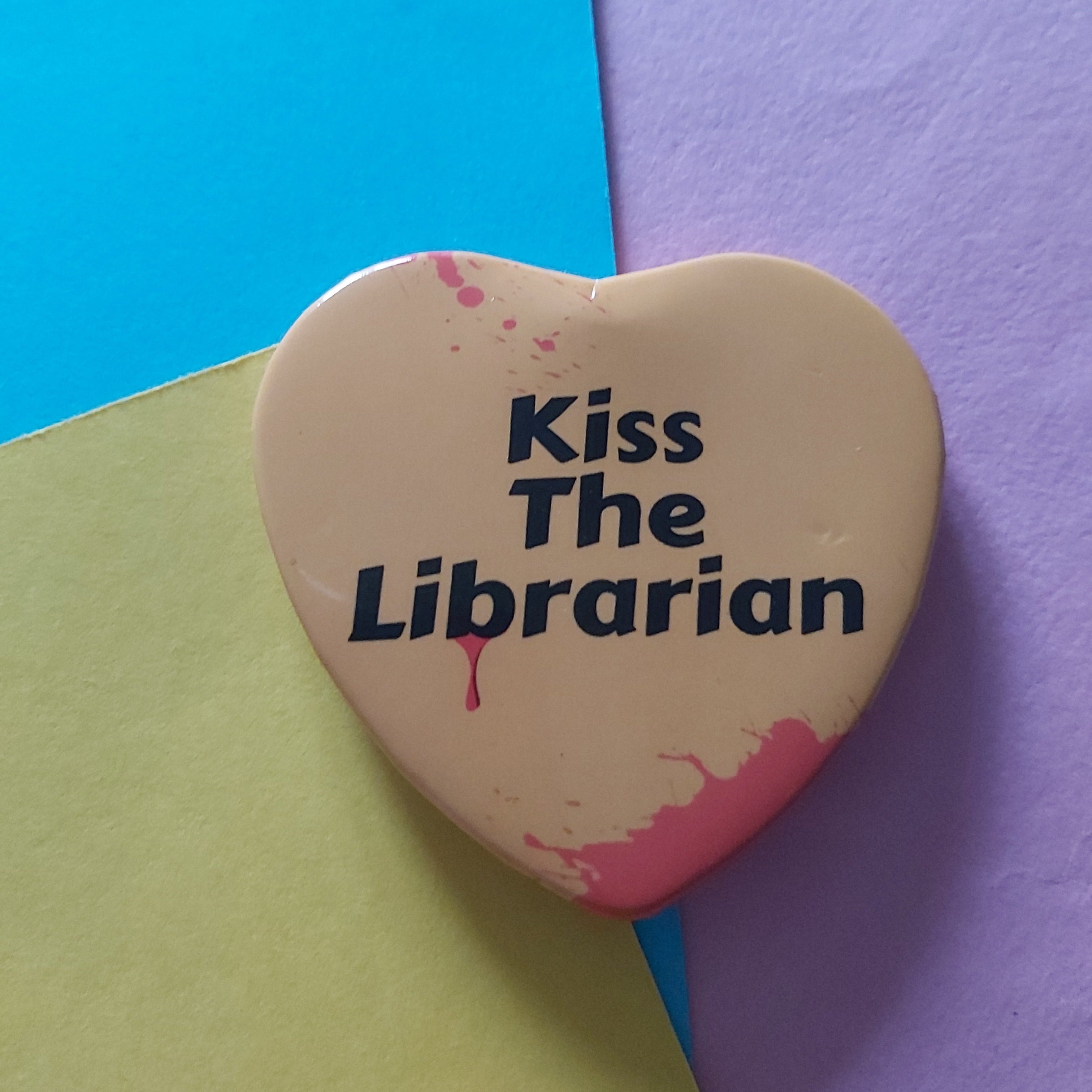 A yellow heart-shaped pin badge with the text 'Kiss The Librarian', placed on a background of three colored paper sheets in blue, purple, and pale yellow-green.