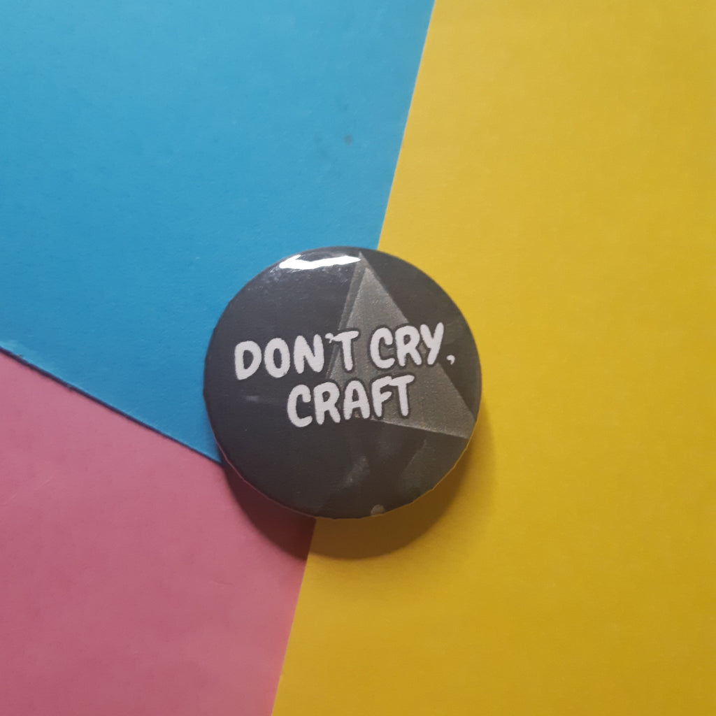 A round pin badge with the text 'DON'T CRY, CRAFT' in white on a dark gray background, placed on a colorful background of blue, yellow, and pink paper.