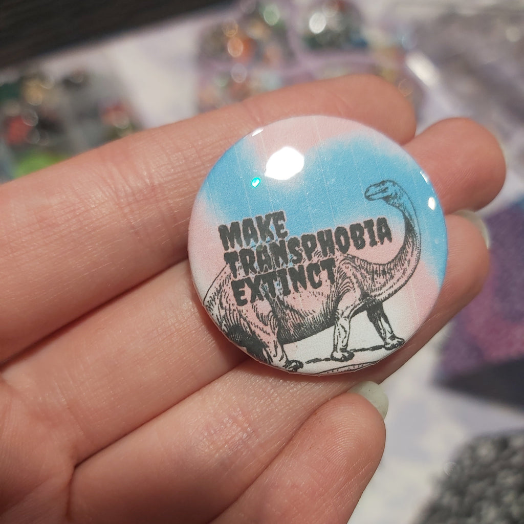 A hand holding a round pin-back button badge with a pastel blue, pink, and white gradient background, featuring a black line drawing of a dinosaur and the text 'MAKE TRANSPHOBIA EXTINCT'.