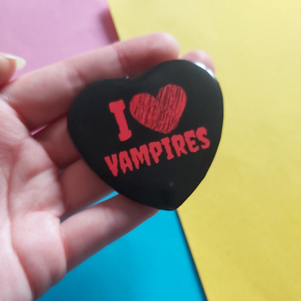 A hand holding a black heart-shaped badge with red text that reads 'I [heart] VAMPIRES' against a colourful background of pink, yellow, and blue paper.