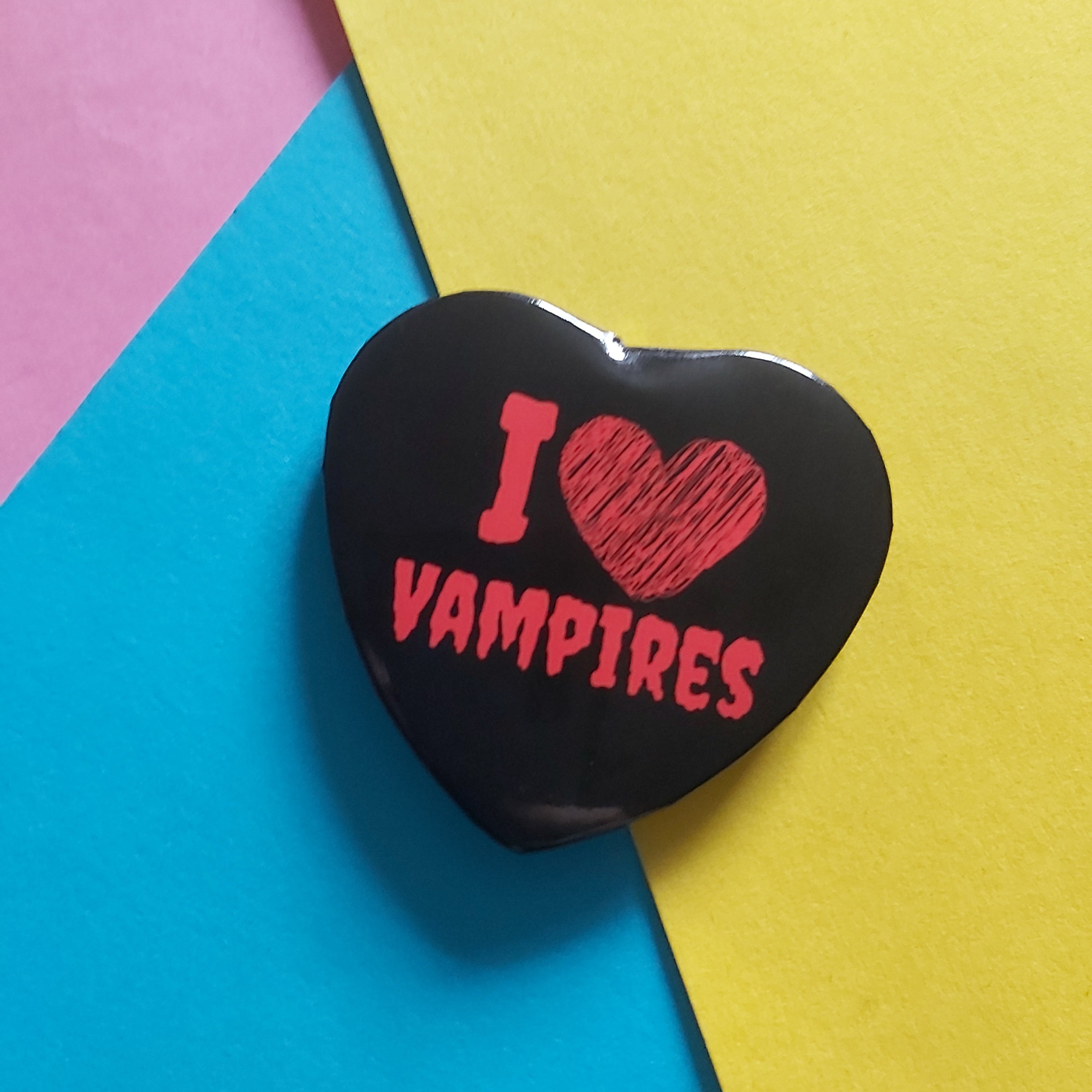 A heart-shaped black badge with red text reading 'I [heart] VAMPIRES' on a background of diagonal pink, blue, and yellow paper sheets.