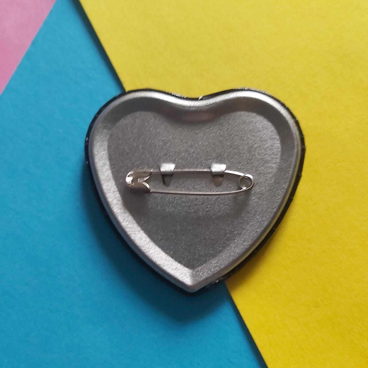 A heart-shaped silver safety pin pinback button shown from the back, resting on a diagonally split background of bright blue and yellow paper.