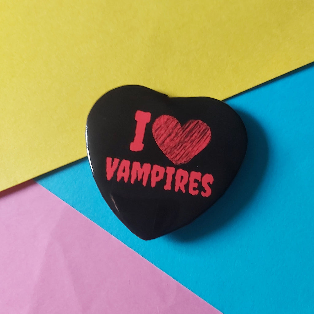 A heart-shaped black pin with red text reading 'I [heart] VAMPIRES' placed on a colorful background of yellow, blue, and pink paper.