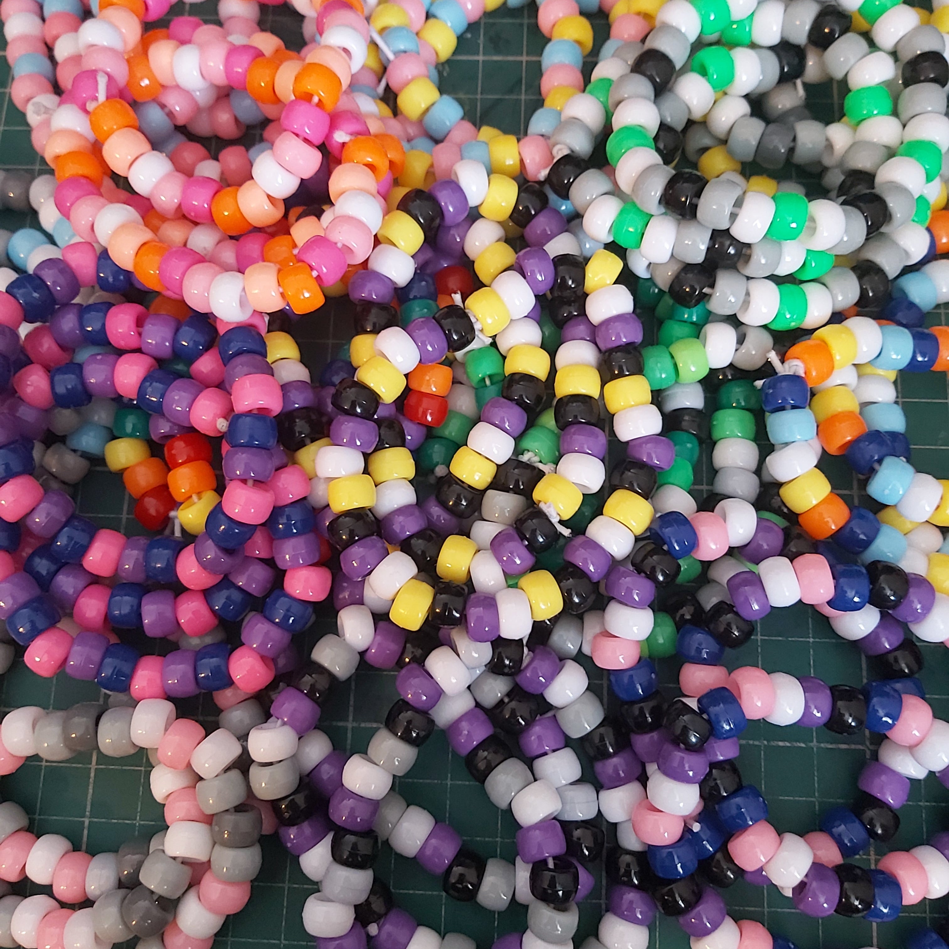 A large assortment of colourful beaded bracelets laid out on a green cutting mat. The beads come in many different colours, including pink, orange, yellow, purple, blue, green, black, and white, arranged in various patterns.