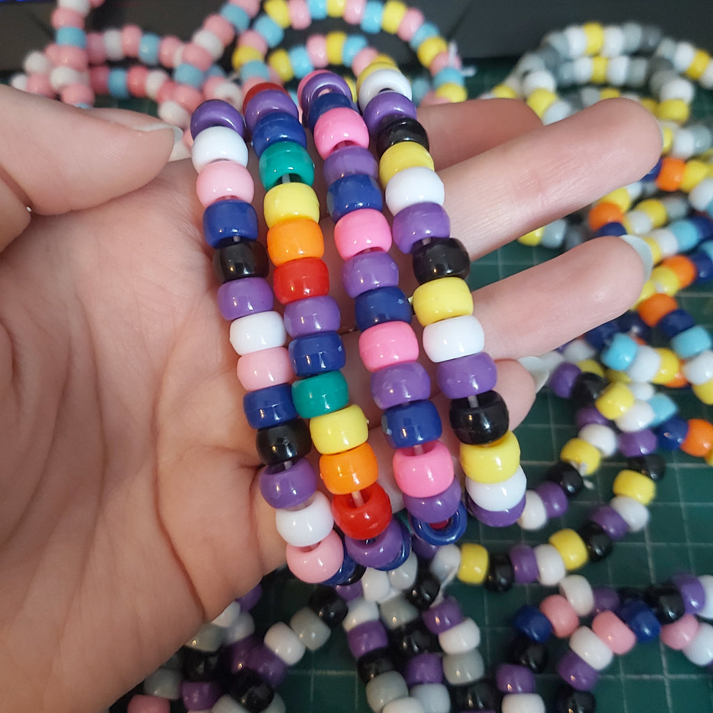 A hand holding four colourful beaded bracelets in multiple colours, including pink, purple, blue, orange, yellow, white, and black, with more of the same bracelets scattered in the background on a green cutting mat.