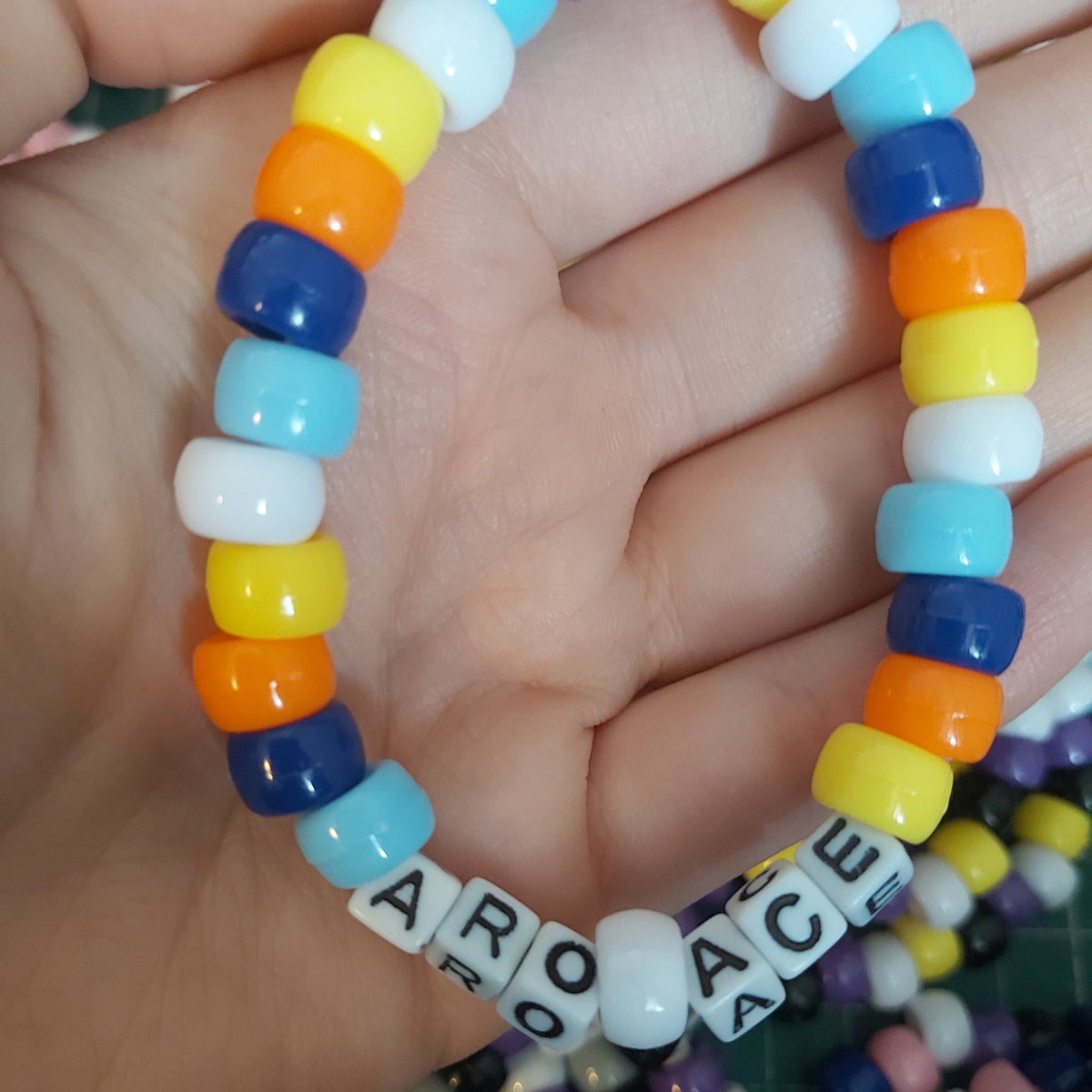 A handmade beaded bracelet held in a hand, featuring colourful round beads in blue, yellow, orange, white, and light blue, with white letter beads spelling 'ARO ACE'.