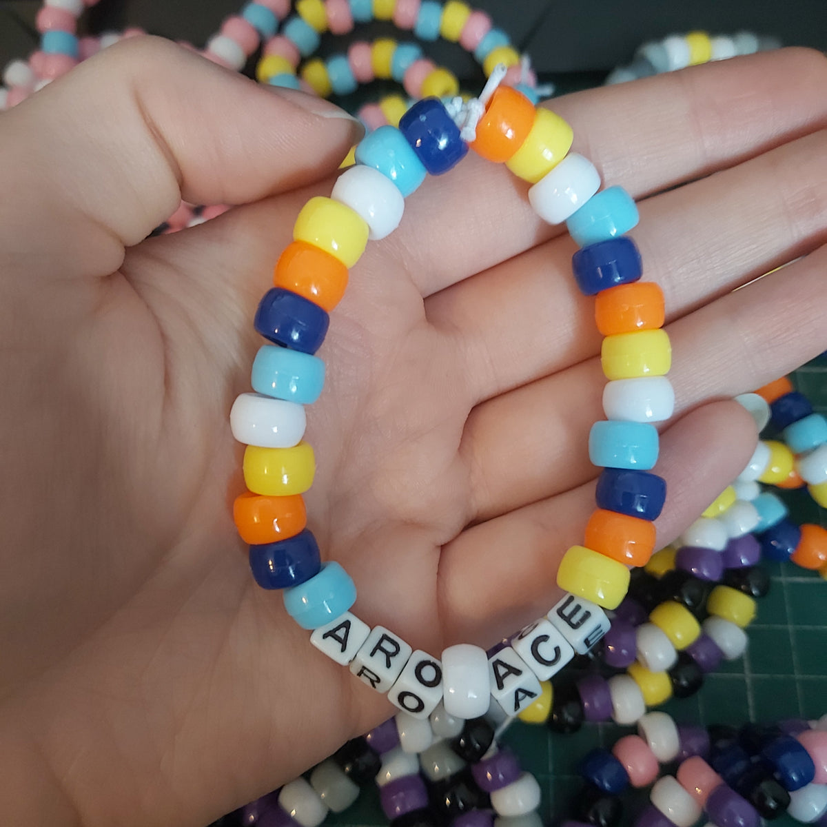 A hand holding a beaded stretch bracelet made of colourful round beads in blue, orange, yellow, white, and light blue, with letter tiles spelling out 'ARO ACE'.