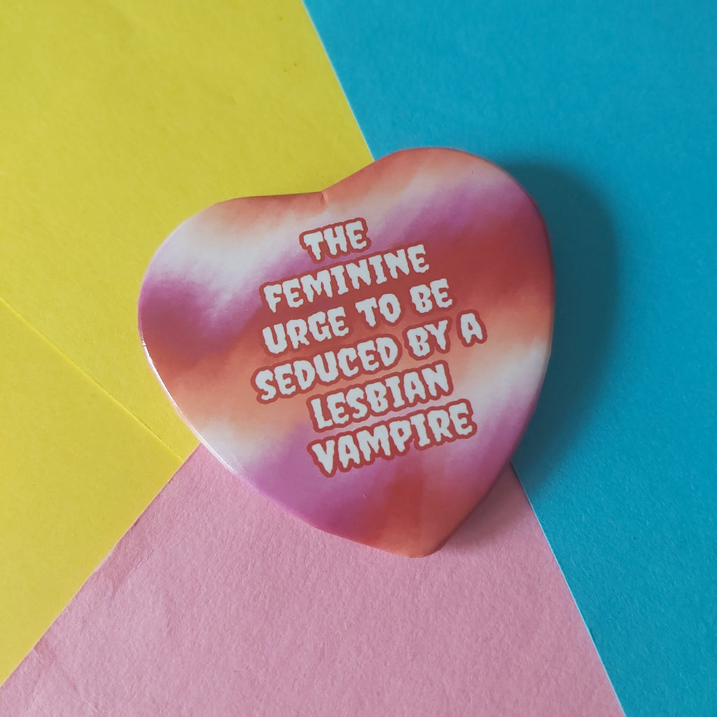 A heart-shaped pinback button with a pink, orange, and white tie-dye pattern and the text 'THE FEMININE URGE TO BE SEDUCED BY A LESBIAN VAMPIRE' in white bubble letters with a pink outline, set against a background of yellow, blue, and pink paper.