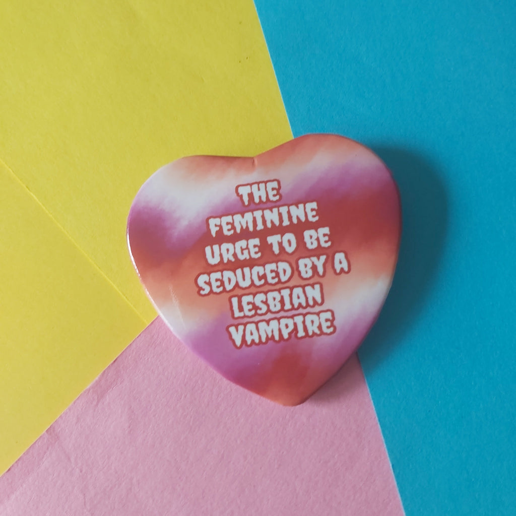 A heart-shaped badge with a pink, orange, and white tie-dye pattern featuring the text 'THE FEMININE URGE TO BE SEDUCED BY A LESBIAN VAMPIRE' in white with a pink outline, placed on a diagonally split yellow, blue, and pink background.