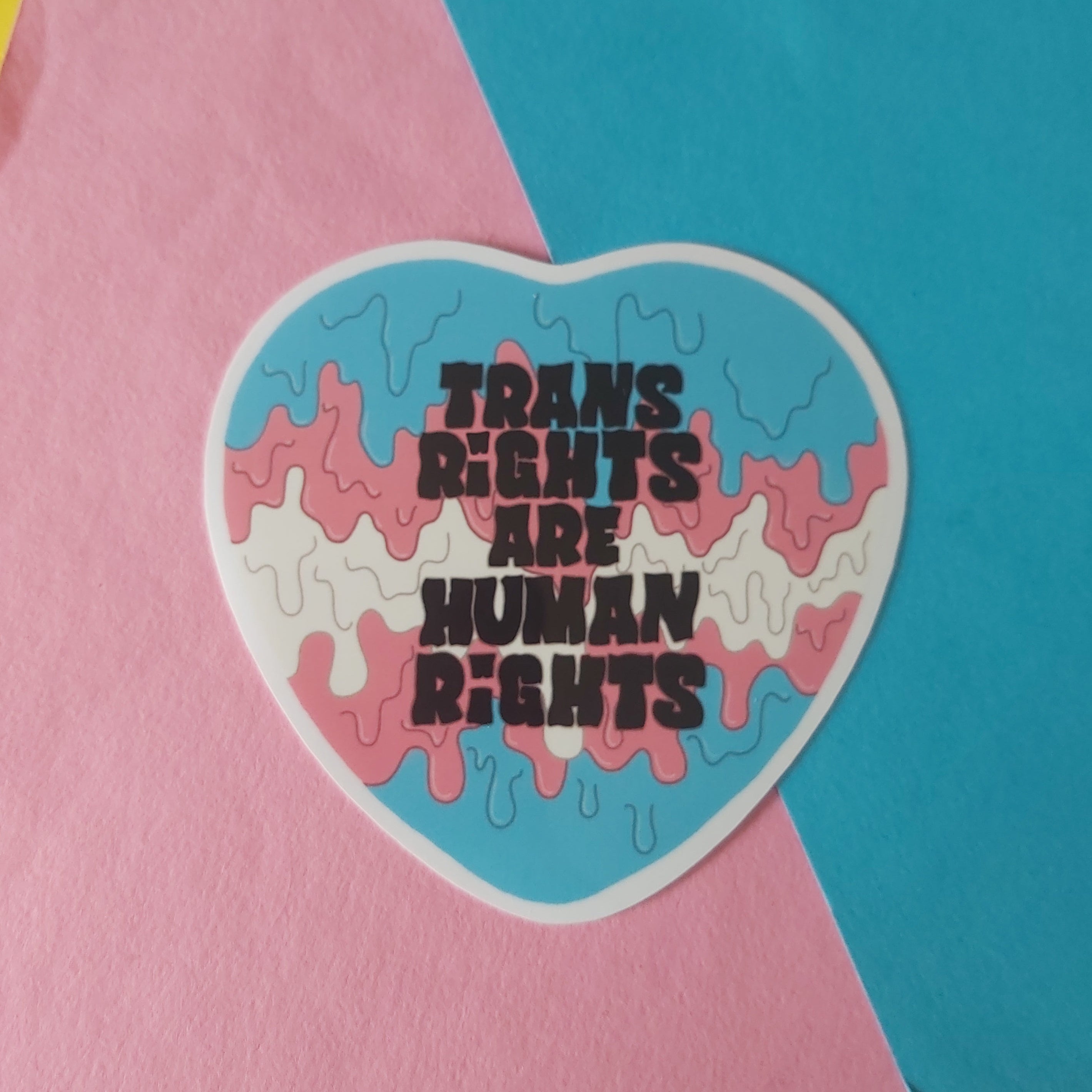 A heart-shaped sticker with the text 'TRANS RIGHTS ARE HUMAN RIGHTS' in black over a pink, blue, and white dripping design, placed on a background of pink, blue, and yellow paper.