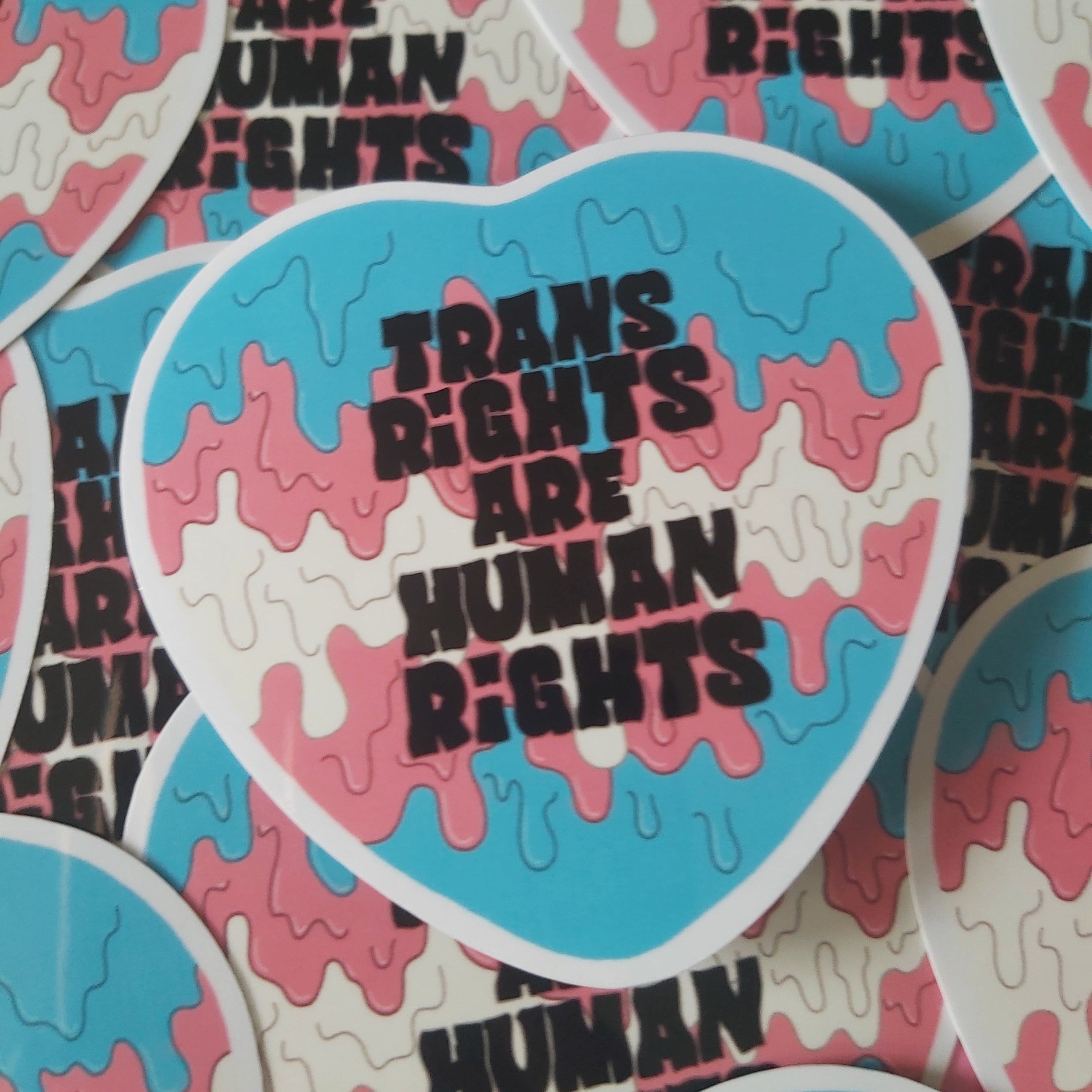 Heart-shaped stickers with a pink, blue, and white dripping paint pattern and the text 'TRANS RIGHTS ARE HUMAN RIGHTS' in black, shown overlapping on a white background.