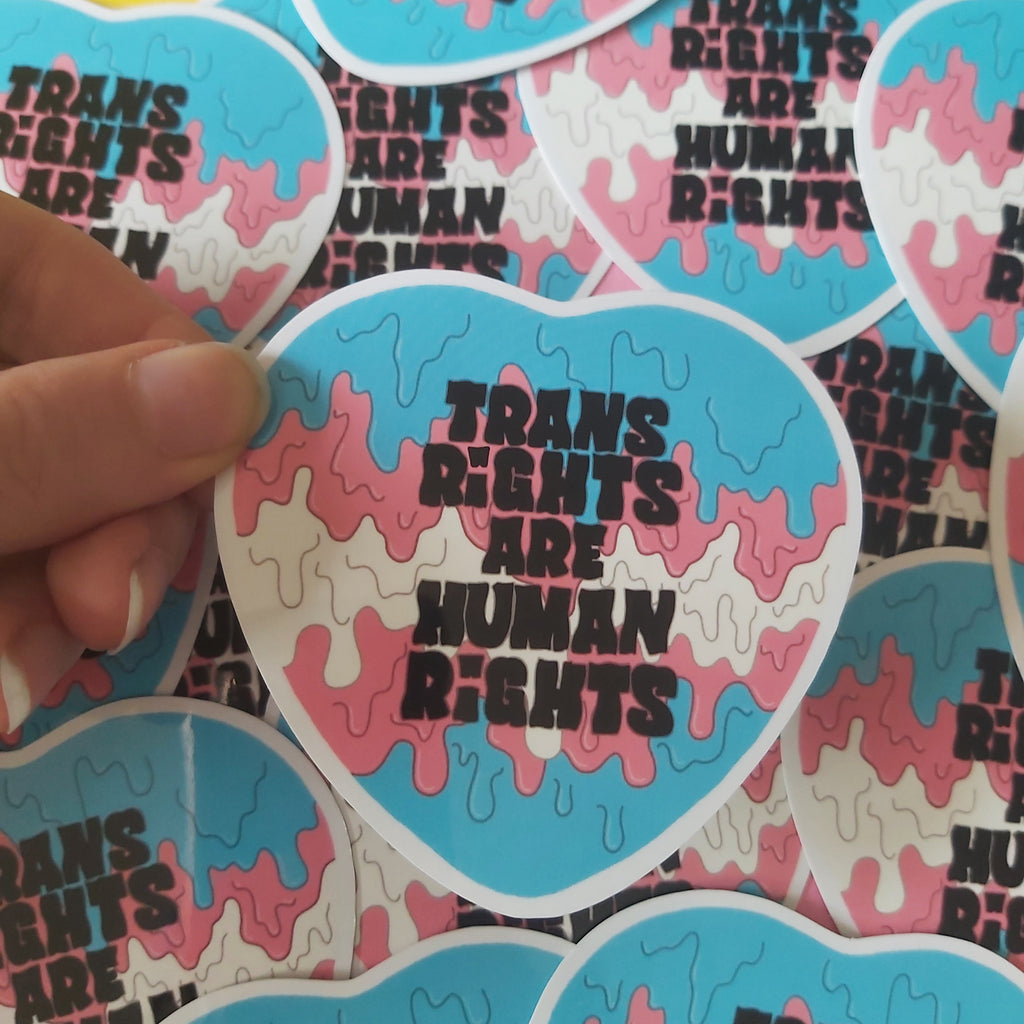 A hand holds a heart-shaped sticker with a pink and light blue paint drip and the text 'TRANS RIGHTS ARE HUMAN RIGHTS' in black. More identical stickers are scattered in the background.