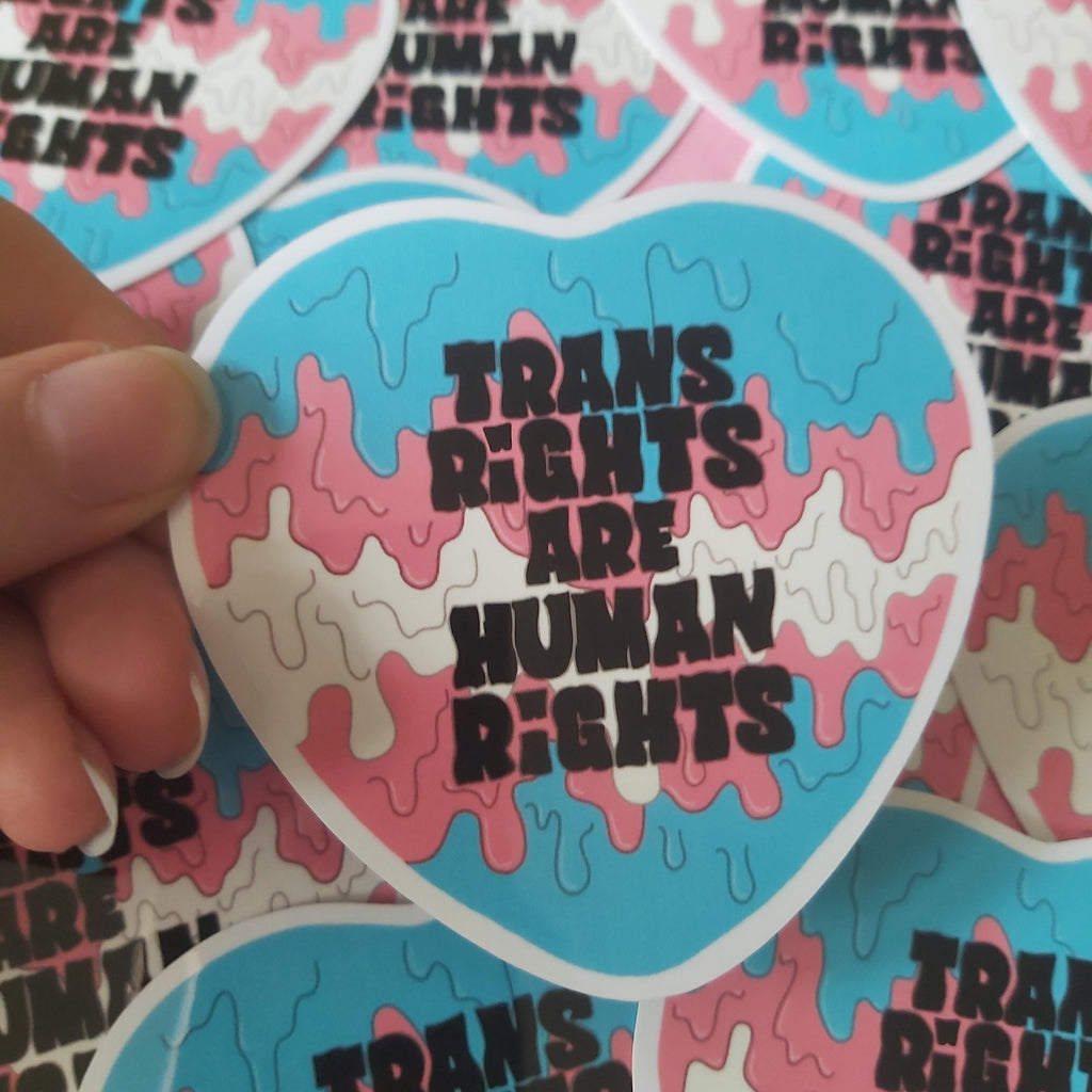 A close-up of a hand holding a heart-shaped sticker with the text 'TRANS RIGHTS ARE HUMAN RIGHTS' in black, with more identical stickers scattered in the background.