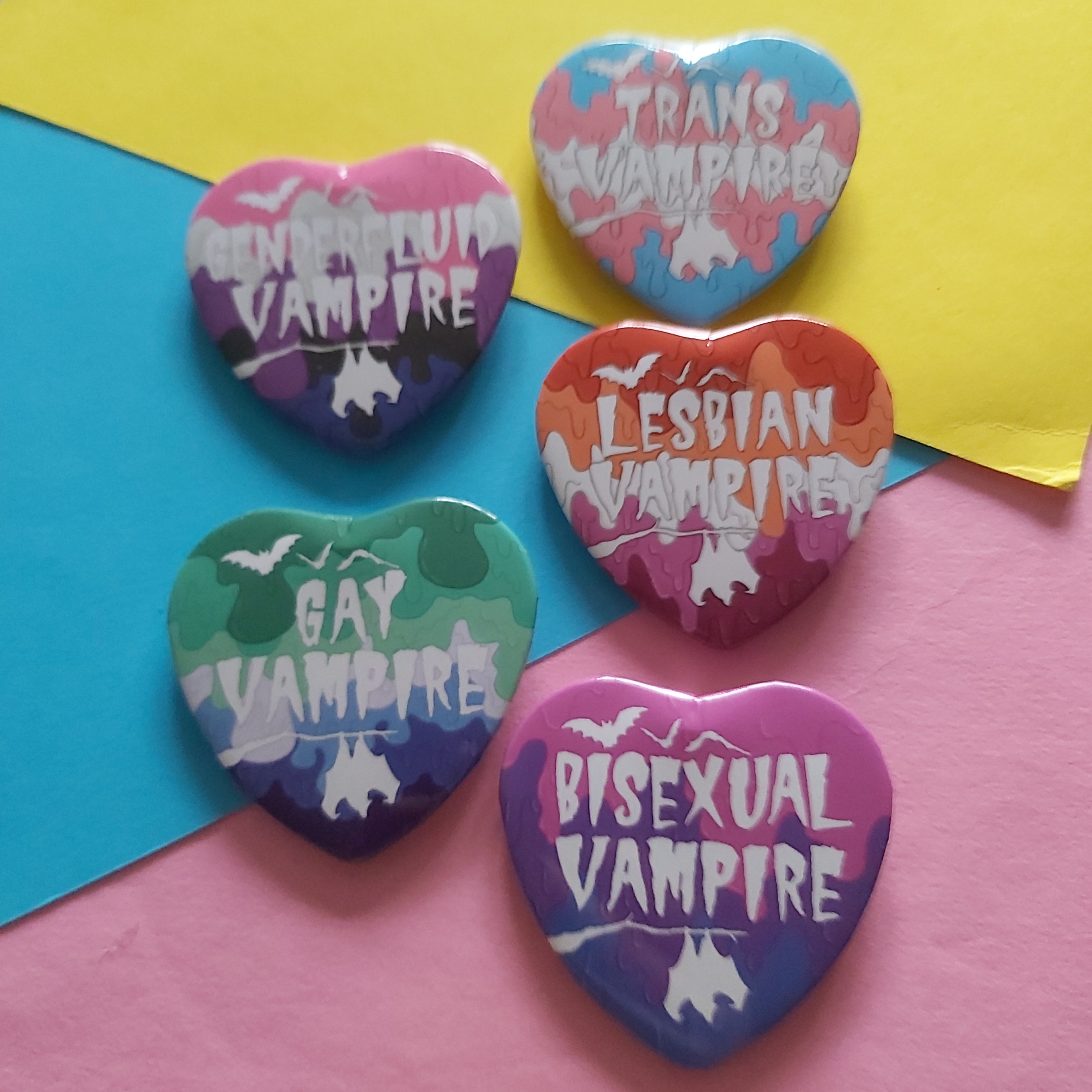 Five heart-shaped pins with white text reading LGBTQ+ vampire themes, arranged on a colourful background of pink, yellow, and blue paper.