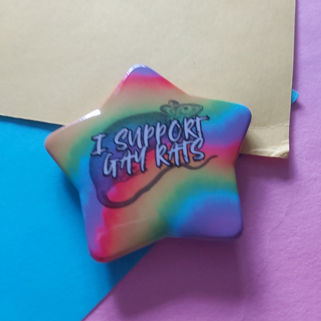 A star-shaped pinback button featuring a rainbow tie-dye pattern and the text 'I SUPPORT GAY RATS' in stylized lettering, displayed on a background of blue, purple, and beige paper.