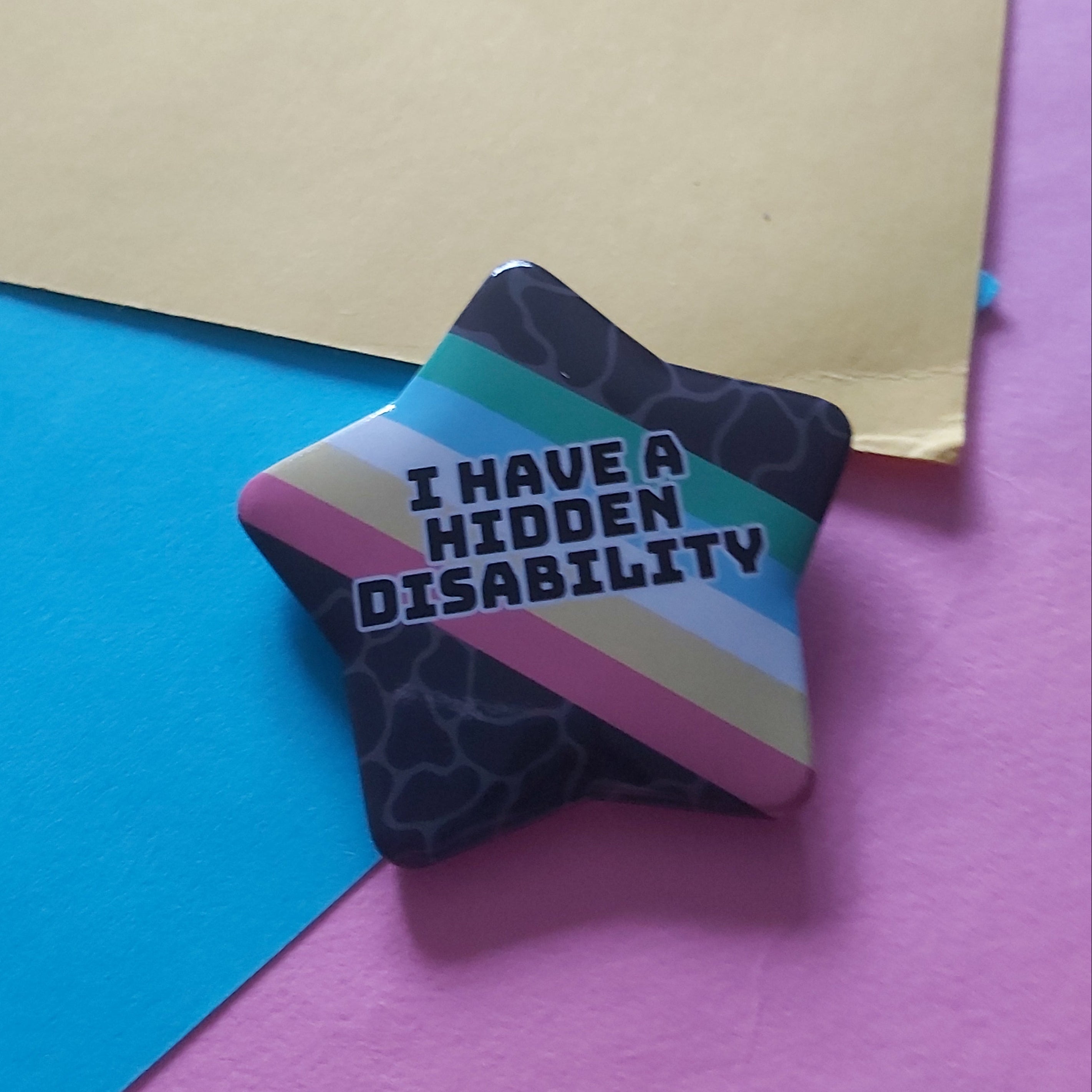 A star-shaped pin badge with the text 'I HAVE A HIDDEN DISABILITY' in white letters, featuring a Disability Pride flag design and a dark patterned background, resting on colourful paper sheets.