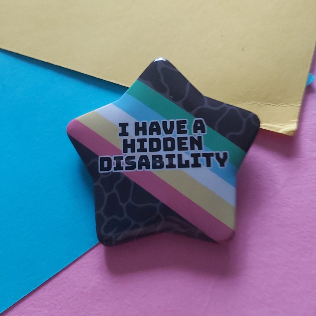 A star-shaped pin badge with the text 'I HAVE A HIDDEN DISABILITY' in white letters, featuring a dark patterned background and diagonal stripes in pink, teal, light blue, and beige, resting on a split pink and blue background with a yellow sheet of paper.