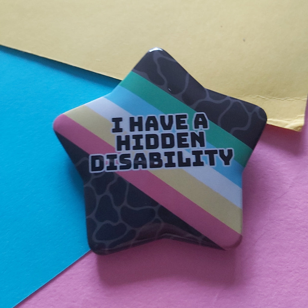 A star-shaped pin badge with the text 'I HAVE A HIDDEN DISABILITY' in white letters, featuring diagonal stripes in teal, light blue, pink, beige, and white over a dark patterned background, displayed on a colorful paper backdrop.