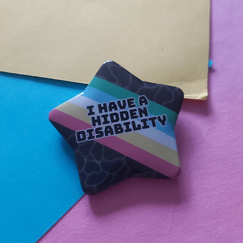 A star-shaped pin badge with the text 'I HAVE A HIDDEN DISABILITY' in white letters, set against diagonal stripes of green, light blue, white, yellow, red and a dark patterned background. The pin is displayed on a colourful background of blue, pink, and yellow paper.