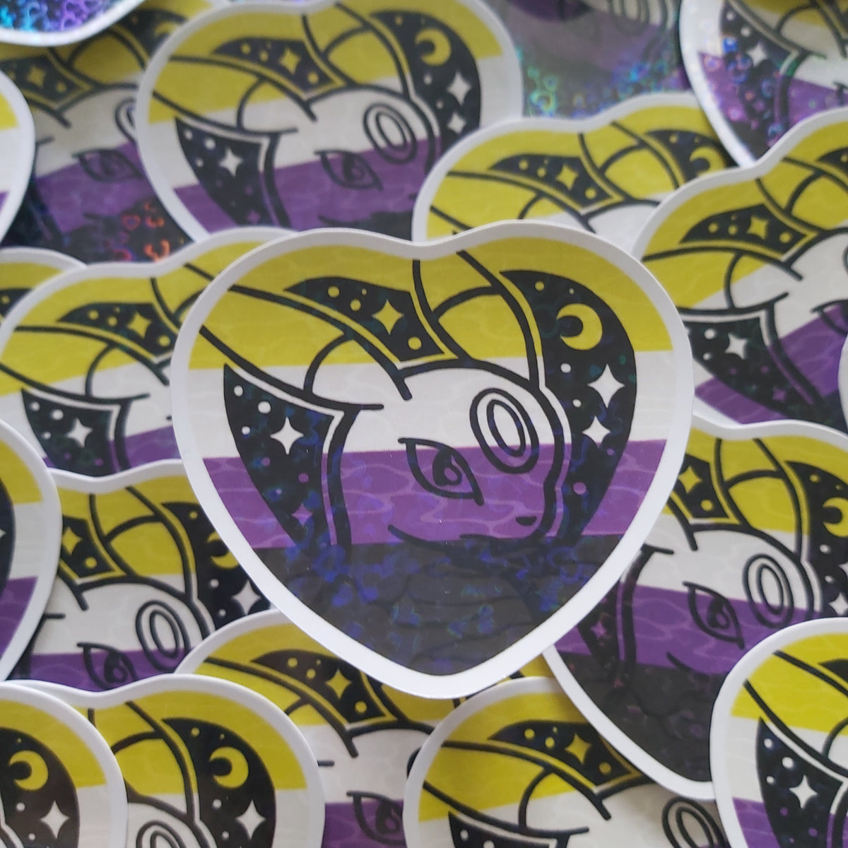 A close-up of multiple heart-shaped stickers scattered on a surface. The central sticker shows a stylized pokemon over a purple, black, and white striped background with yellow accents.