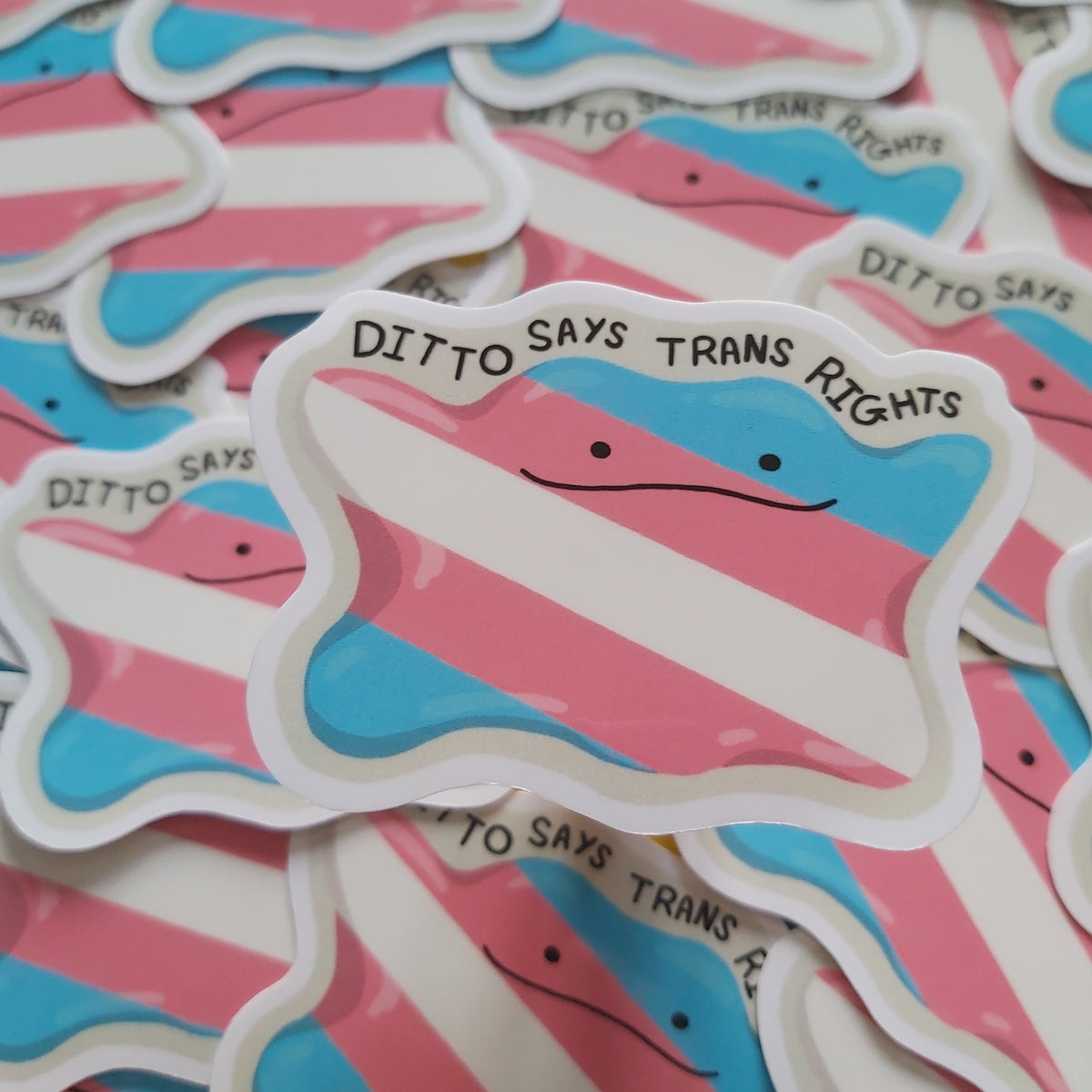 A close-up of multiple die-cut stickers featuring a simple face with pink, light blue, and white horizontal stripes and the text 'DITTO SAYS TRANS RIGHTS' on a white border. The stickers are scattered across the frame, with one centered in focus.